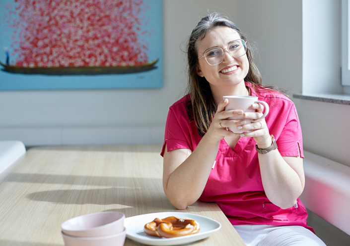 Eine Mitarbeiterin sitzt an einem Tisch mit einer Brezel auf einem Teller und mit einer Tasse in der Hand und lächelt freundlich