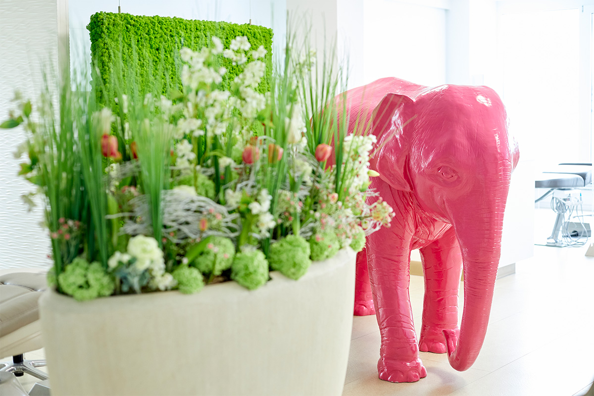 Ein rosa Elefant steht rechts von einem Pflanzenkübel, eine grüne Pflanzenwand steht im Hintergrund